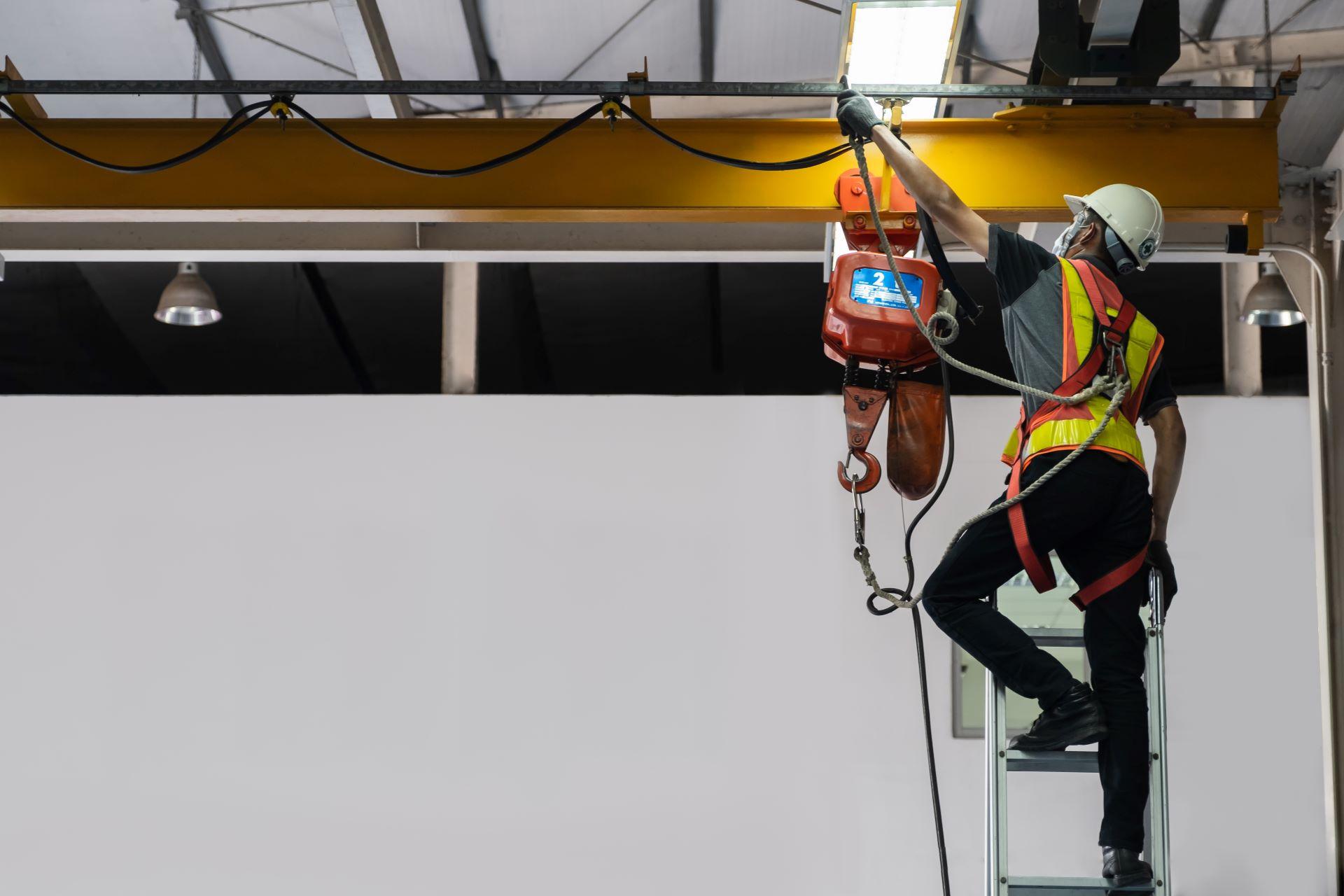 Arbeiter auf Leiter wartet Elektrokettenzug an gelbem Kranträger